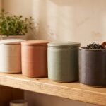 Four ceramic tea canisters in muted earth tones lined up on a wooden kitchen shelf, one open showing dried tea leaves, in warm soft light.
