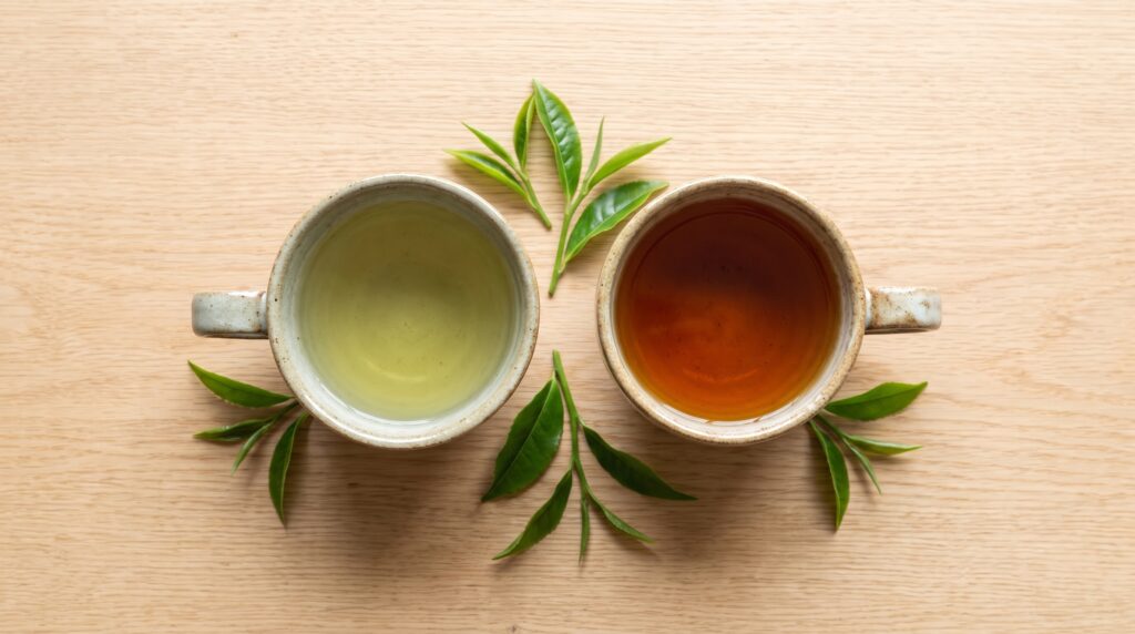Two ceramic cups, one holding pale green tea and one holding deep amber black tea, photographed from above on a wooden table with fresh tea leaves.