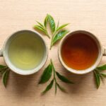 Two ceramic cups, one holding pale green tea and one holding deep amber black tea, photographed from above on a wooden table with fresh tea leaves.