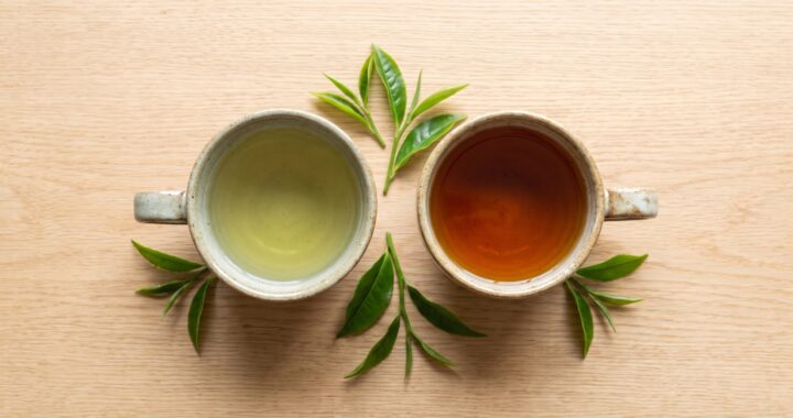Two ceramic cups, one holding pale green tea and one holding deep amber black tea, photographed from above on a wooden table with fresh tea leaves.