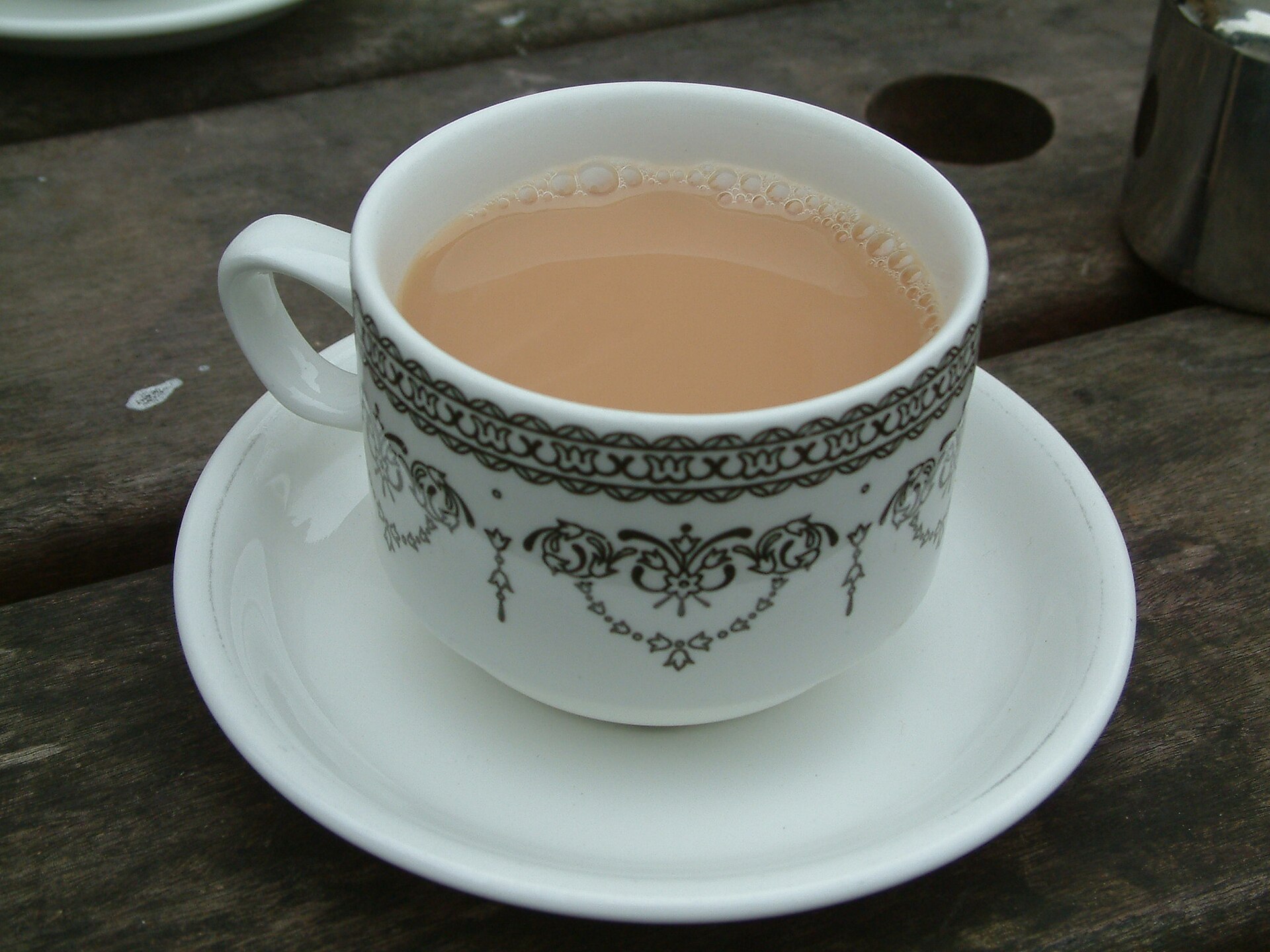 A white china cup and saucer with an ornate black border, filled with pale milky tea, on a weathered wooden table.