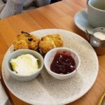 Two fruit scones on a plate with bowls of clotted cream and strawberry jam, beside a cup of tea.