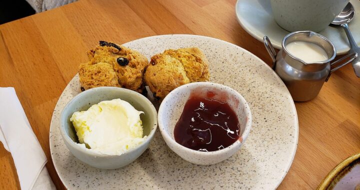 Two fruit scones on a plate with bowls of clotted cream and strawberry jam, beside a cup of tea.