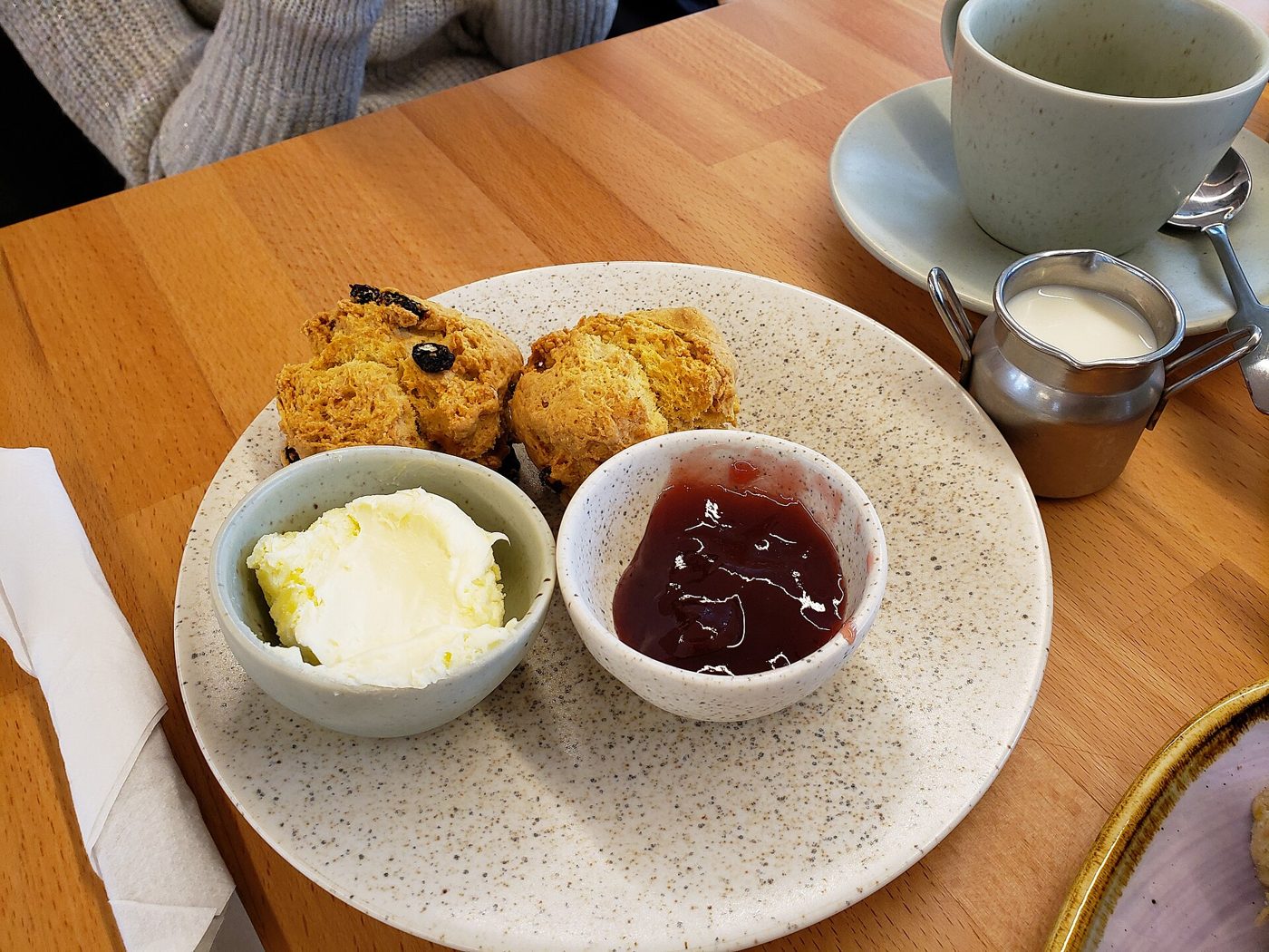 Two fruit scones on a speckled plate with one bowl of clotted cream and one of strawberry jam, beside a cup of tea and a small jug of milk on a wooden café table.