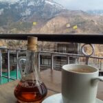 A white mug of hot lemon and ginger tea on a saucer beside a glass bottle of honey, with mountains behind.