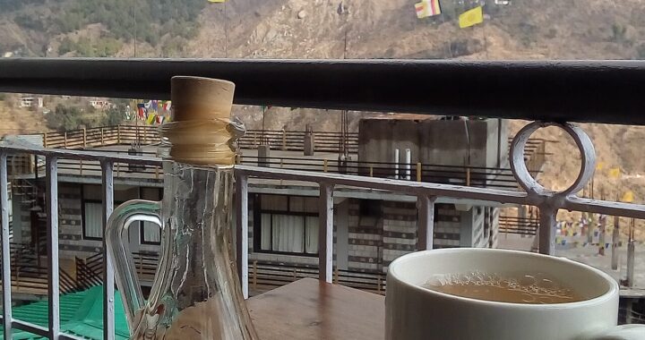 A white mug of hot lemon and ginger tea on a saucer beside a glass bottle of honey, with mountains behind.