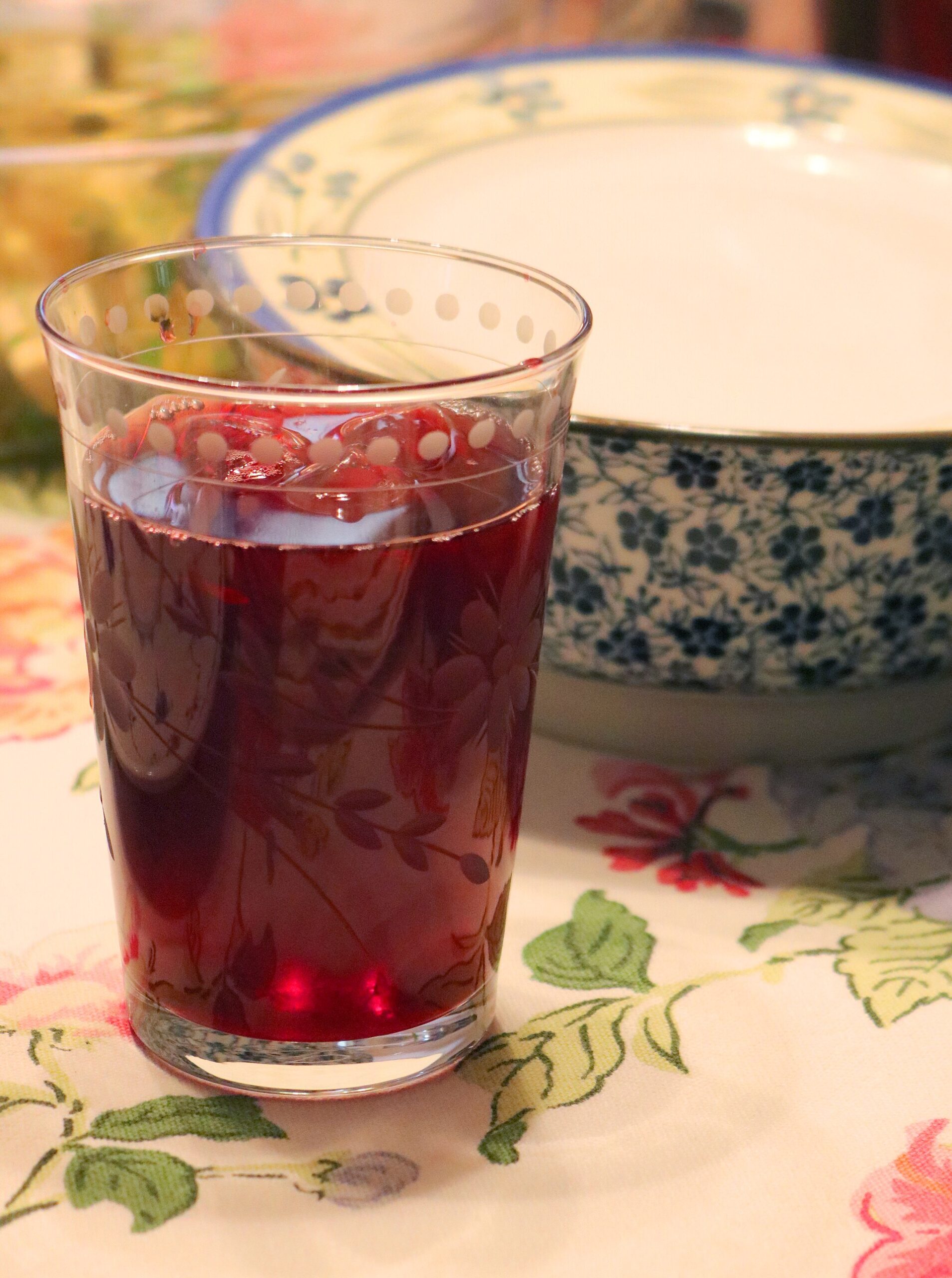 A tall glass of clear dark-red hibiscus tea on a flowered tablecloth, with a blue and white patterned bowl behind it.