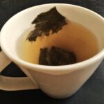 A cream porcelain cup of pale yellow-green tea with a few dark unfurled leaves inside.