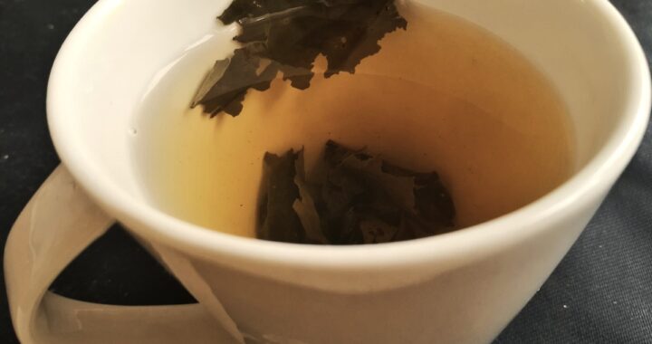 A cream porcelain cup of pale yellow-green tea with a few dark unfurled leaves inside.