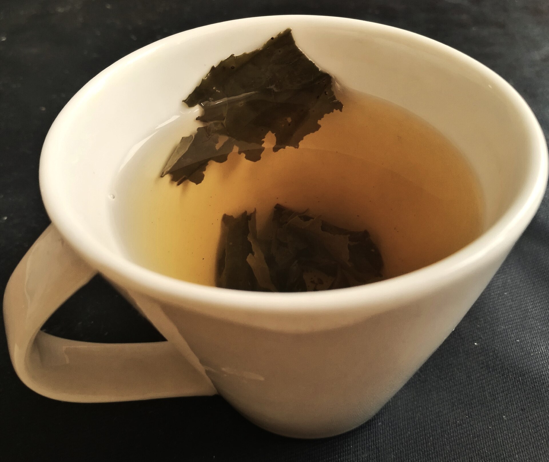 A cream-coloured tilted porcelain cup of pale yellow-green tea with a few dark unfurled tea leaves resting inside.