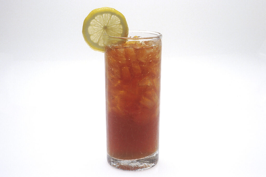 A tall glass of amber iced tea filled with ice, with a lemon slice on the rim, against a white background.