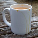 A white mug of milky tea with drips down the side on a weathered wooden table.