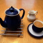 A dark blue teapot on a trivet beside a small white milk jug and a blue cup of milky tea.