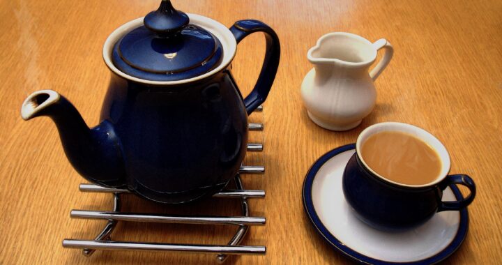 A dark blue teapot on a trivet beside a small white milk jug and a blue cup of milky tea.