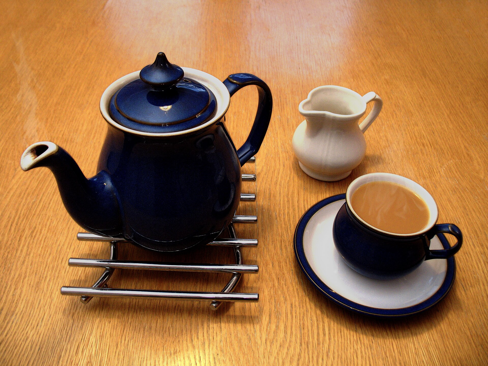 A dark blue teapot on a metal trivet beside a small white milk jug and a blue cup of milky tea on a saucer, on a wooden table.