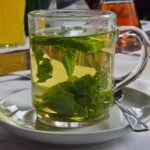 A clear glass mug of pale green-gold tea packed with fresh peppermint leaves, on a saucer.