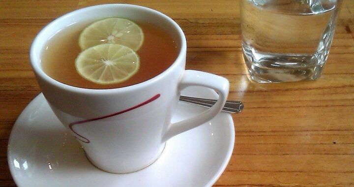 A white cup of tea with two lemon slices on a saucer beside a glass of water on a wooden table.