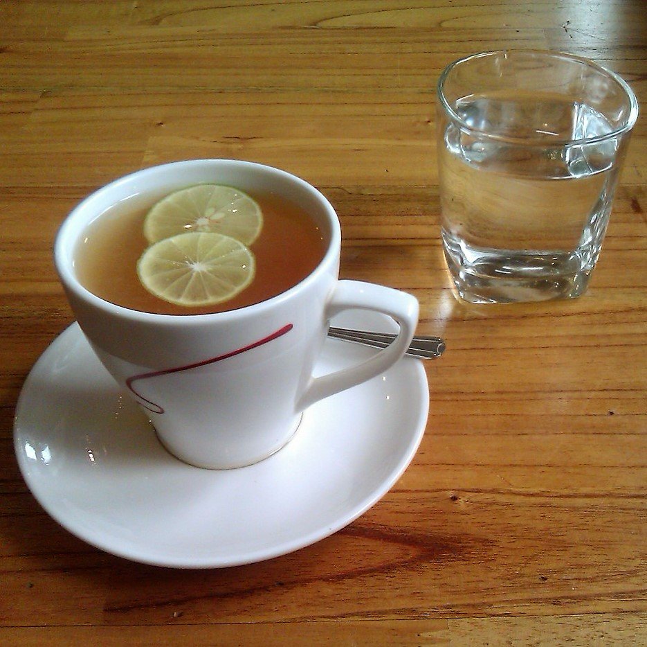 A white cup of tea with two lemon slices on a saucer beside a glass of water on a wooden table.