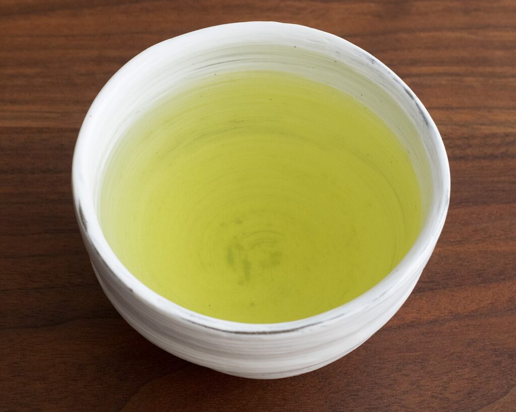 A white ceramic cup of pale yellow-green sencha tea on a dark wooden table, seen from above.