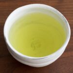 A white ceramic cup of pale yellow-green sencha tea on a dark wooden table, seen from above.