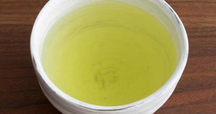A white ceramic cup of pale yellow-green sencha tea on a dark wooden table, seen from above.