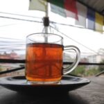 A glass mug of deep amber black tea on a dark saucer, on a table outdoors with prayer flags blurred in the background.