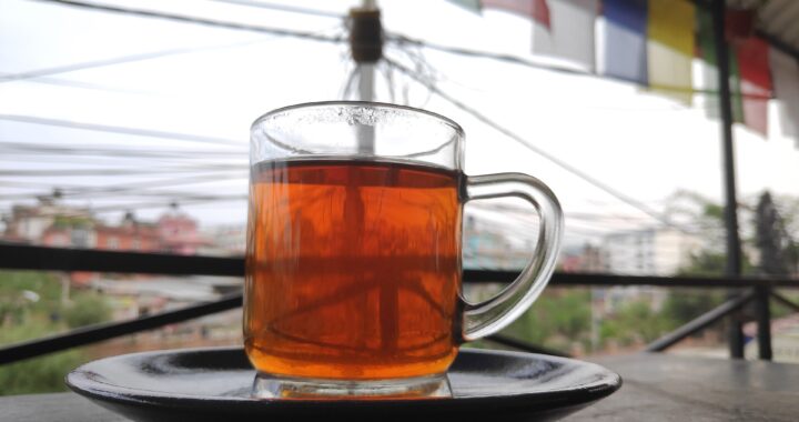 A glass mug of deep amber black tea on a dark saucer, on a table outdoors with prayer flags blurred in the background.