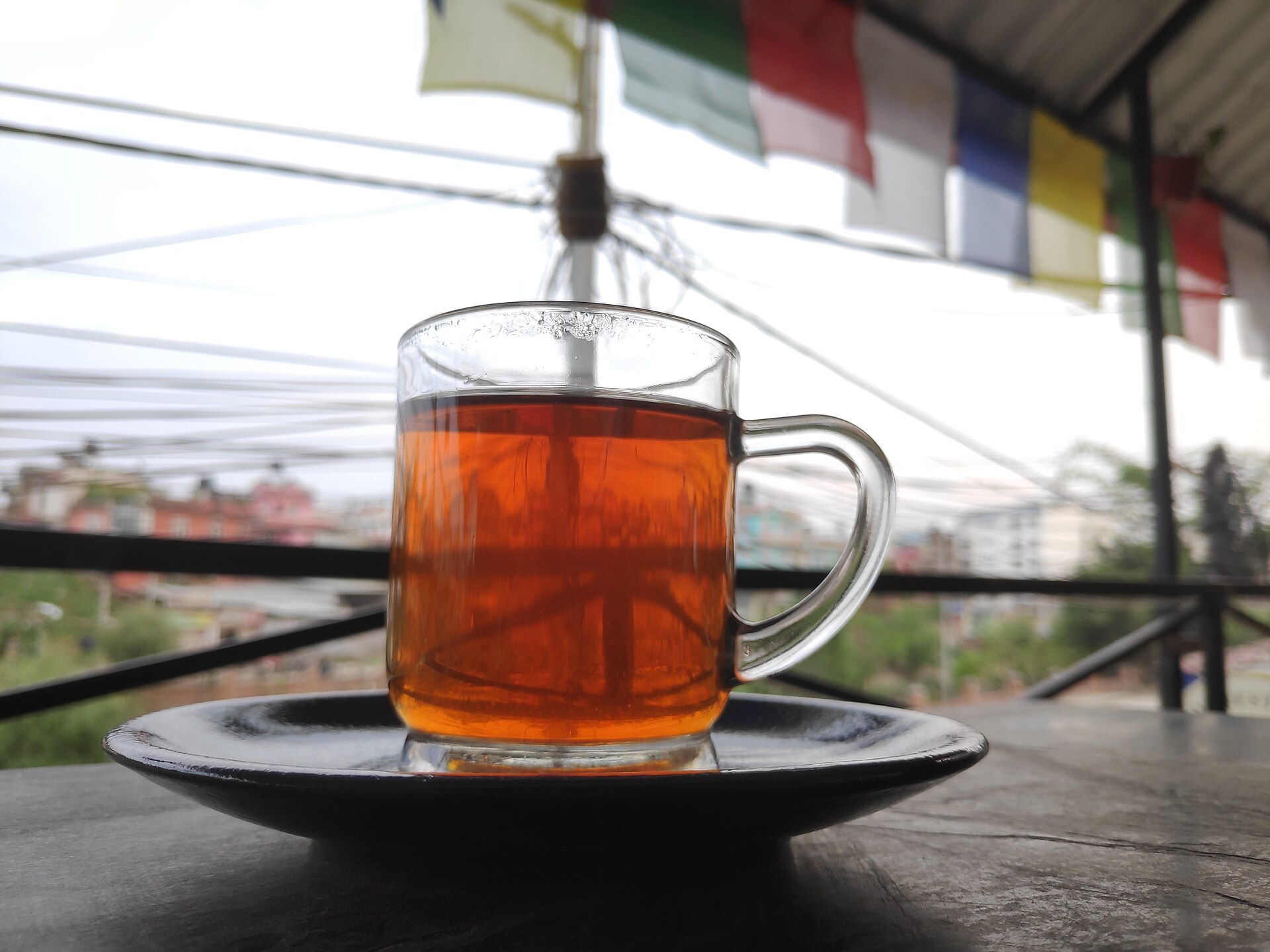 A glass mug of deep amber black tea on a dark saucer, on a table outdoors with prayer flags blurred in the background.