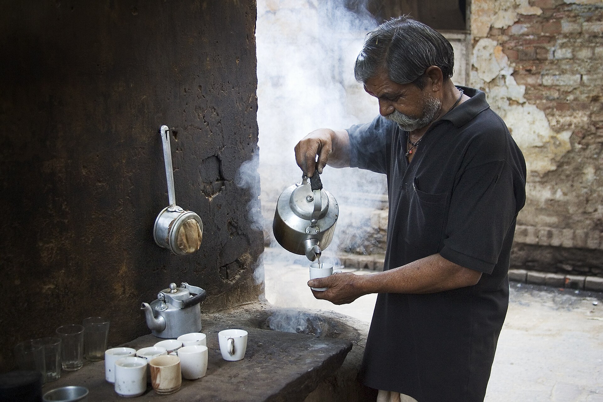 A street vendor in Varanasi pouring tea from a metal kettle into small cups on a stone counter, with steam rising behind him.