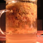 A clear glass mug of pale golden chamomile tea with whole dried chamomile flowers floating at the top, on a wooden table.