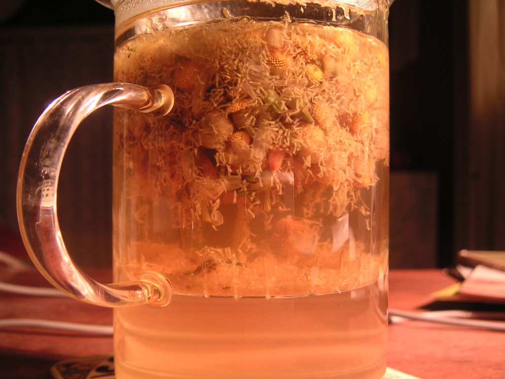 A clear glass mug of pale golden chamomile tea with whole dried chamomile flowers floating at the top, on a wooden table.