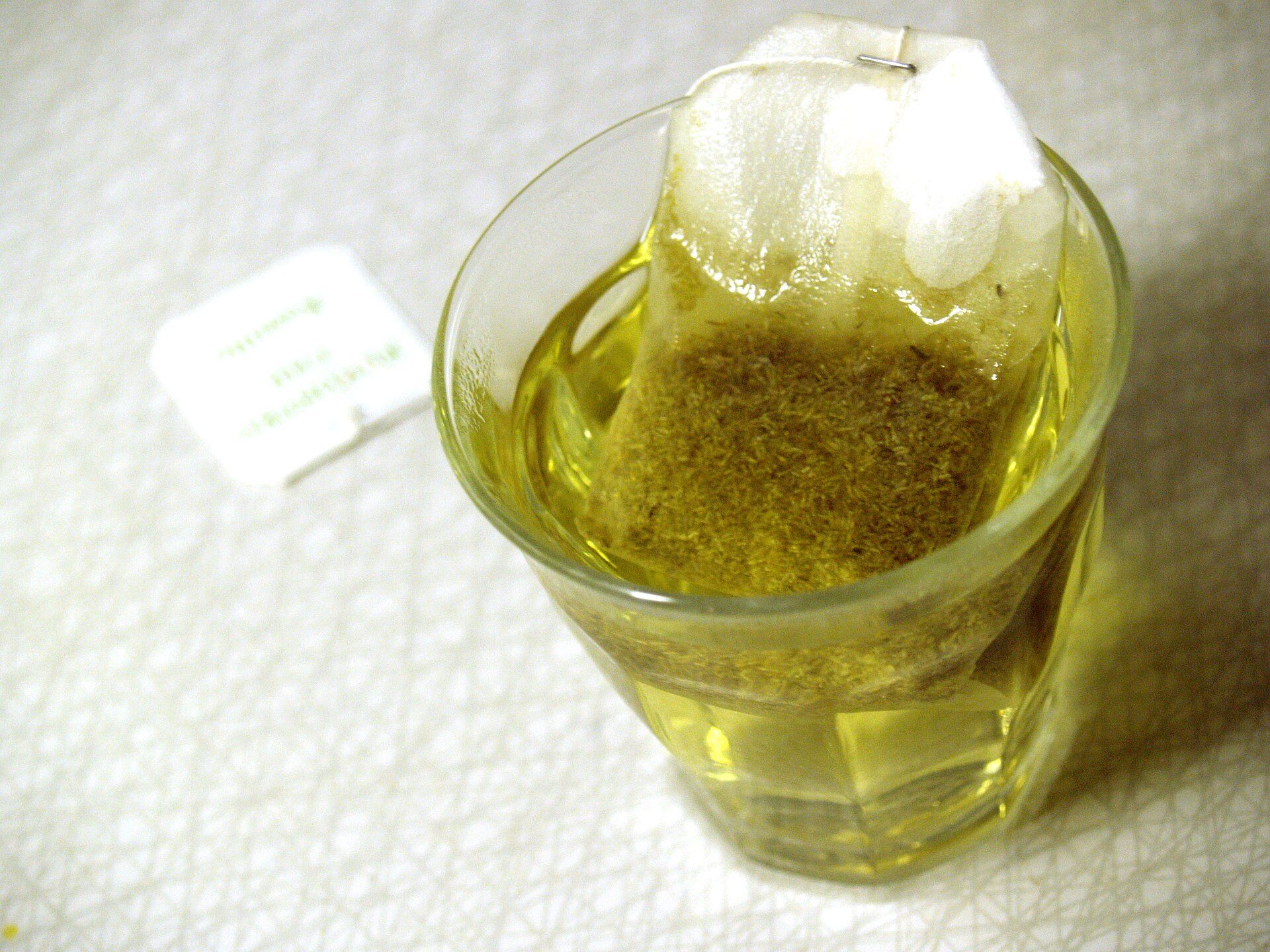 A small glass of pale golden chamomile tea with the teabag still steeping in it, on a white textured cloth.