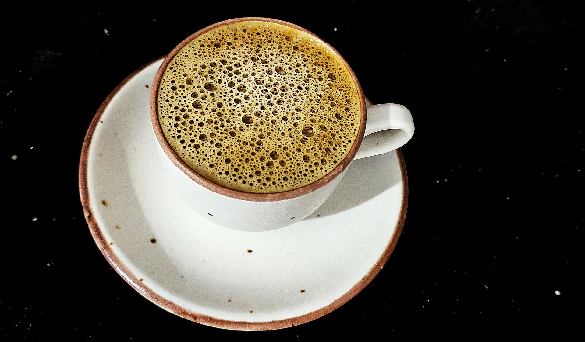 A white cup of frothy coffee on a speckled saucer against a black background.