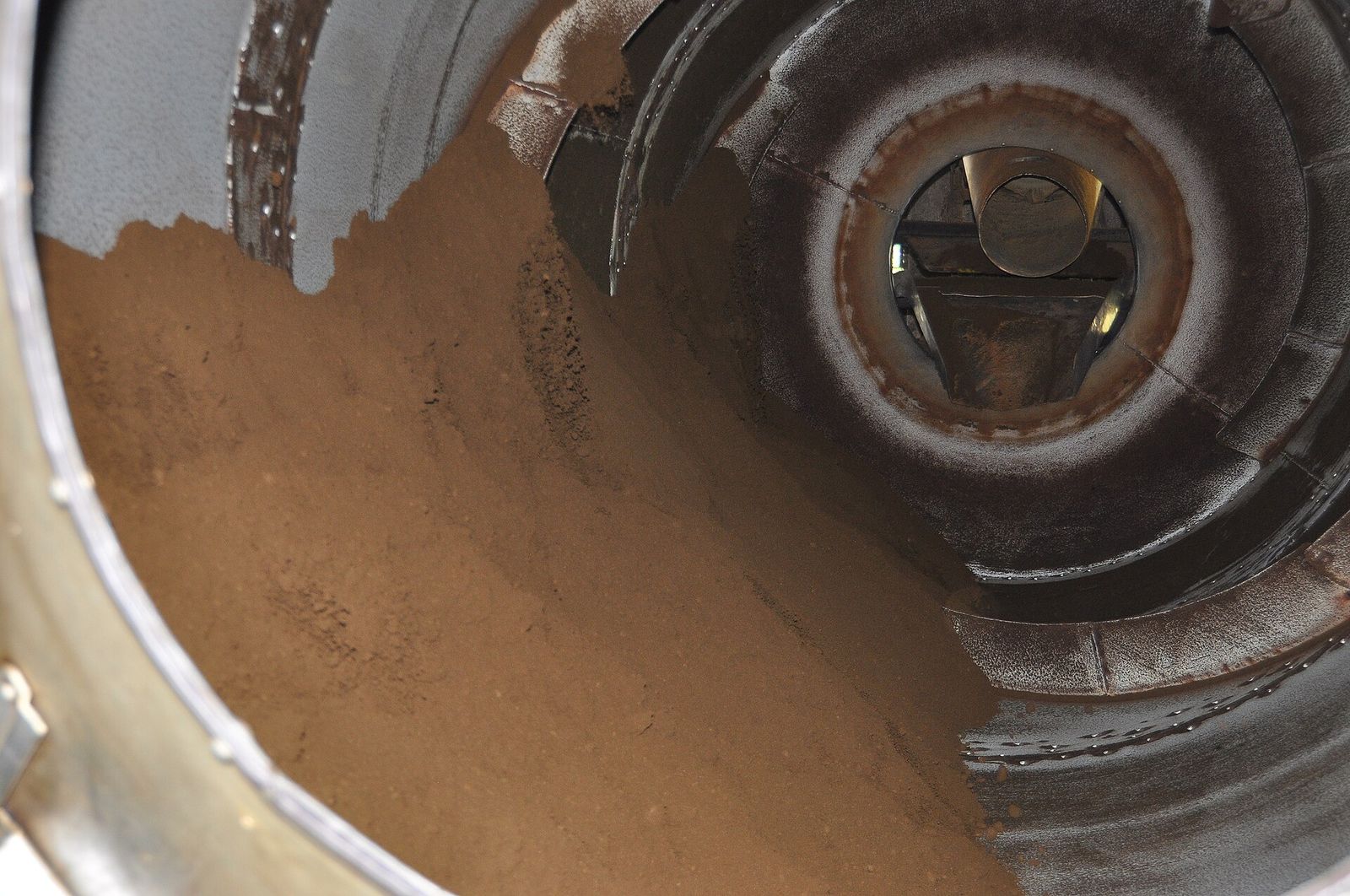 Close-up inside a large metal drying drum half full of fine, reddish-brown CTC tea granules.