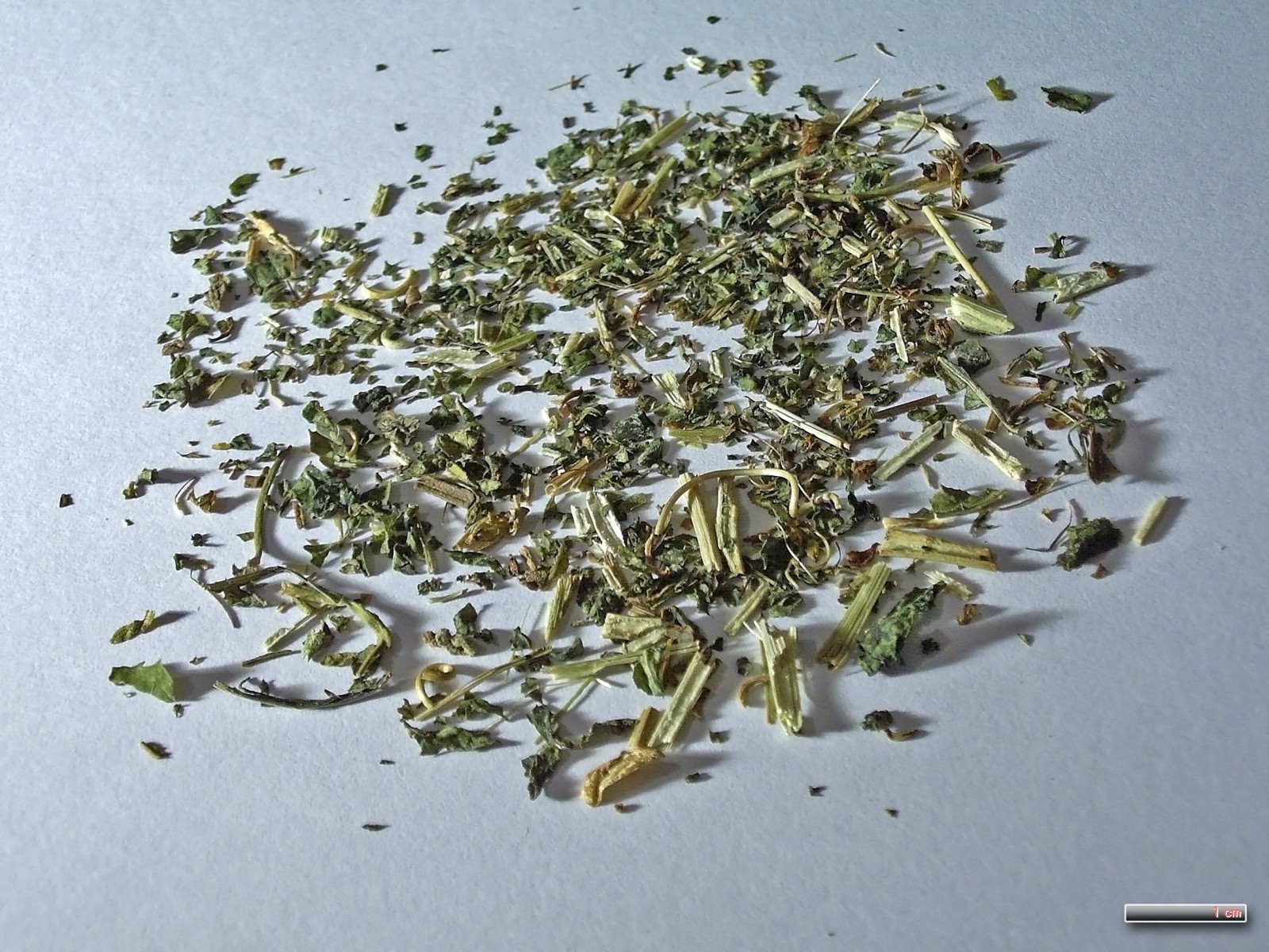 A small heap of dried, cut passionflower herb on white paper: green leaf fragments, pale stem pieces and a few curled tendrils.