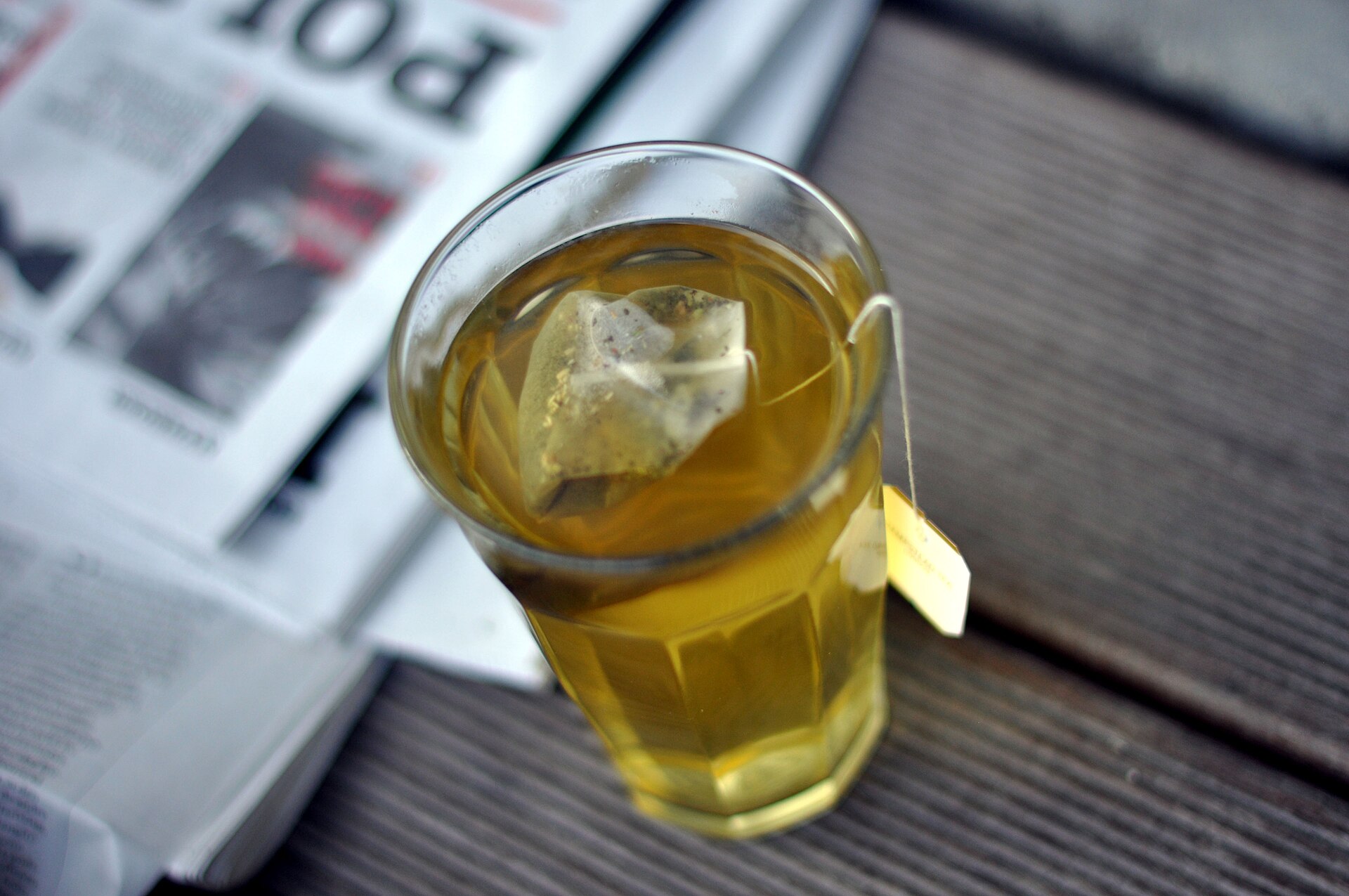 A glass of golden fennel tea with the teabag in it, standing on a newspaper on a dark wooden table.