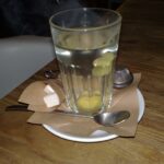 A tall glass of pale ginger tea with fresh ginger slices, on a saucer on a wooden table.