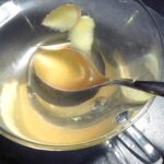 A glass cup of pale golden ginger tea with slices of fresh ginger root and a teaspoon, seen from above on a dark table.