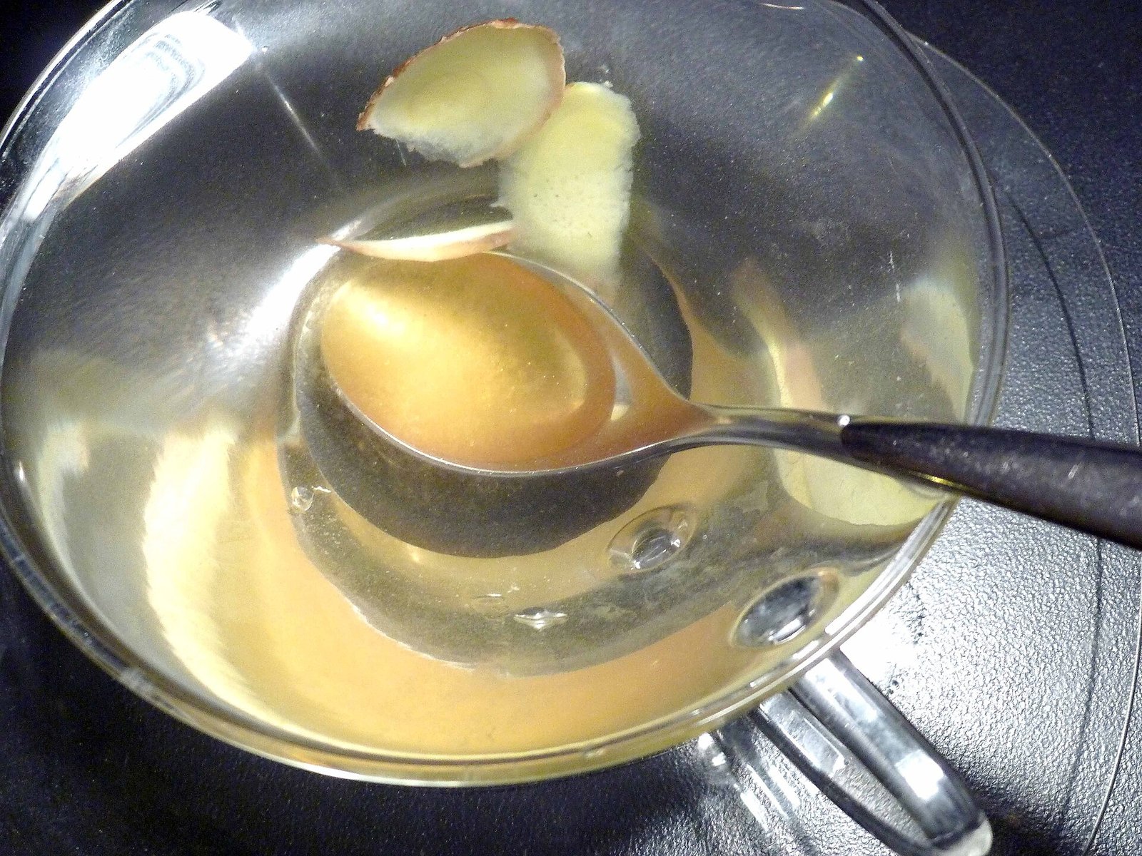 A glass cup of pale golden ginger tea with slices of fresh ginger root and a teaspoon, seen from above on a dark table.