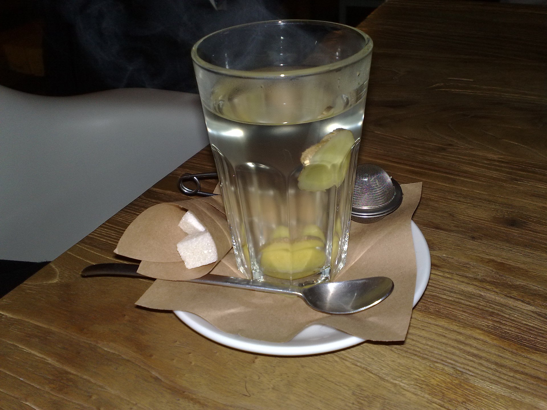 A tall glass of pale ginger tea with slices of fresh ginger at the bottom, on a saucer with a spoon and sugar cubes, on a wooden table.