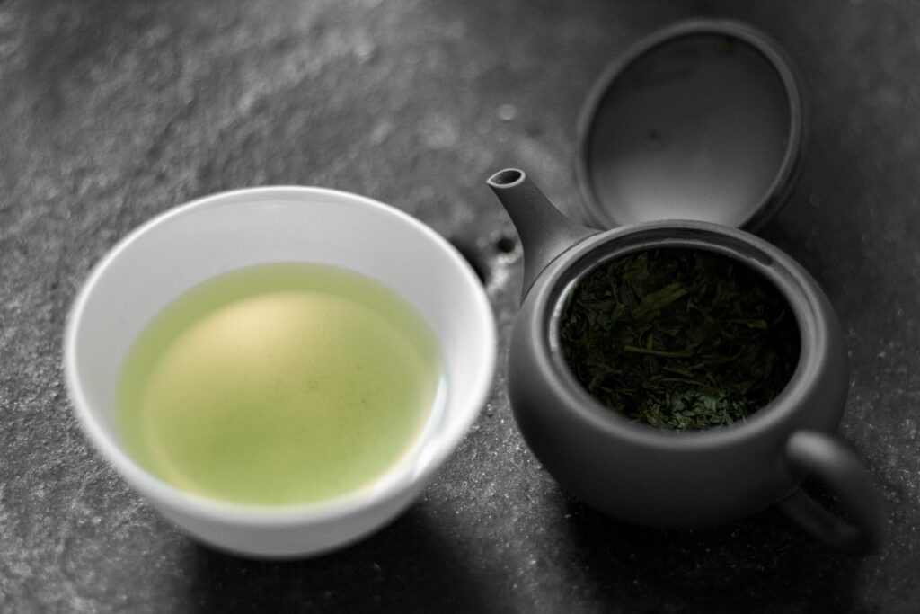 A small white cup of pale green gyokuro tea next to a dark Japanese teapot holding wet tea leaves, on a grey stone surface.