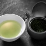 A small white cup of pale green gyokuro tea next to a dark Japanese teapot holding wet tea leaves, on a grey stone surface.
