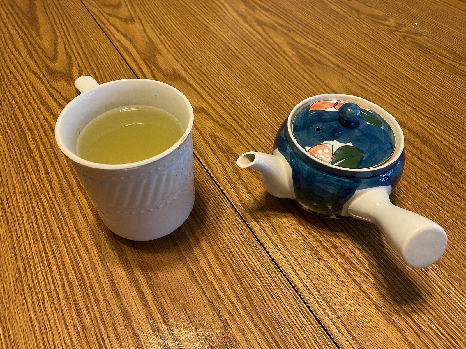 A white ceramic mug of pale yellow-green gyokuro tea on a wooden table, next to a small blue and white side-handled kyusu teapot painted with leaves and orange fruit.