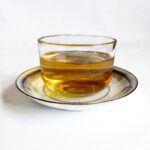 A clear glass cup of golden herbal tea on a gold-rimmed saucer against a white background.