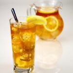 A tall glass of iced tea with ice cubes, a lemon wedge and a spoon, in front of a glass jug of iced tea with lemon slices, on a white background.