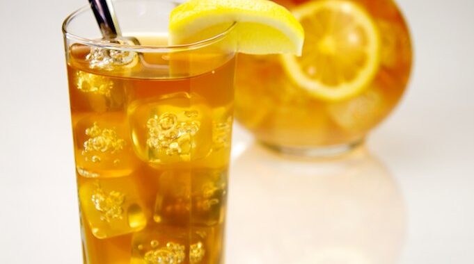 A tall glass of iced tea with ice cubes, a lemon wedge and a spoon, in front of a glass jug of iced tea with lemon slices, on a white background.