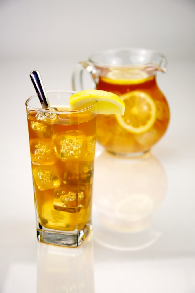 A tall glass of iced tea with ice cubes, a lemon wedge and a spoon, in front of a glass jug of iced tea with lemon slices, on a white background.