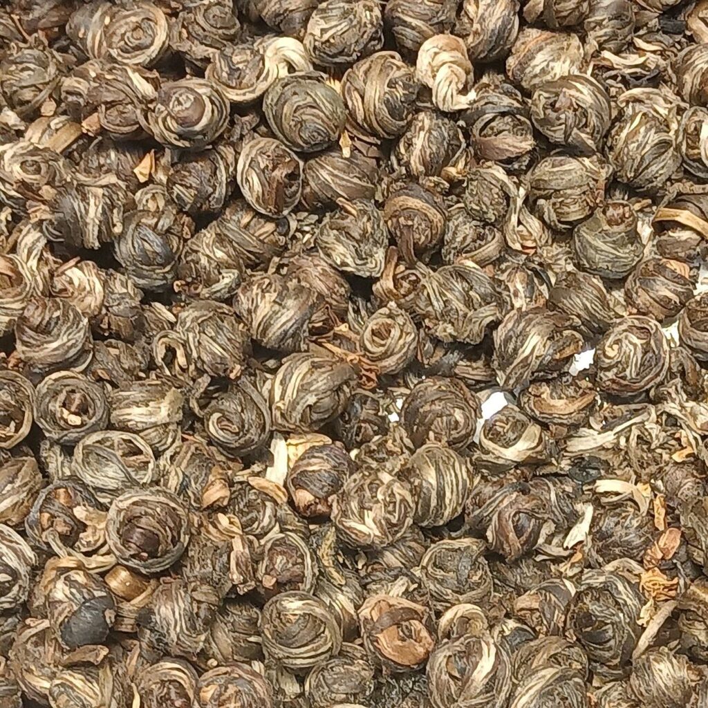 A close view of hundreds of small hand-rolled jasmine tea pearls, pale green and silver with flecks of dried petal.