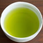 A white ceramic cup filled to near the brim with bright yellow-green sencha, seen from above on a dark wooden table.