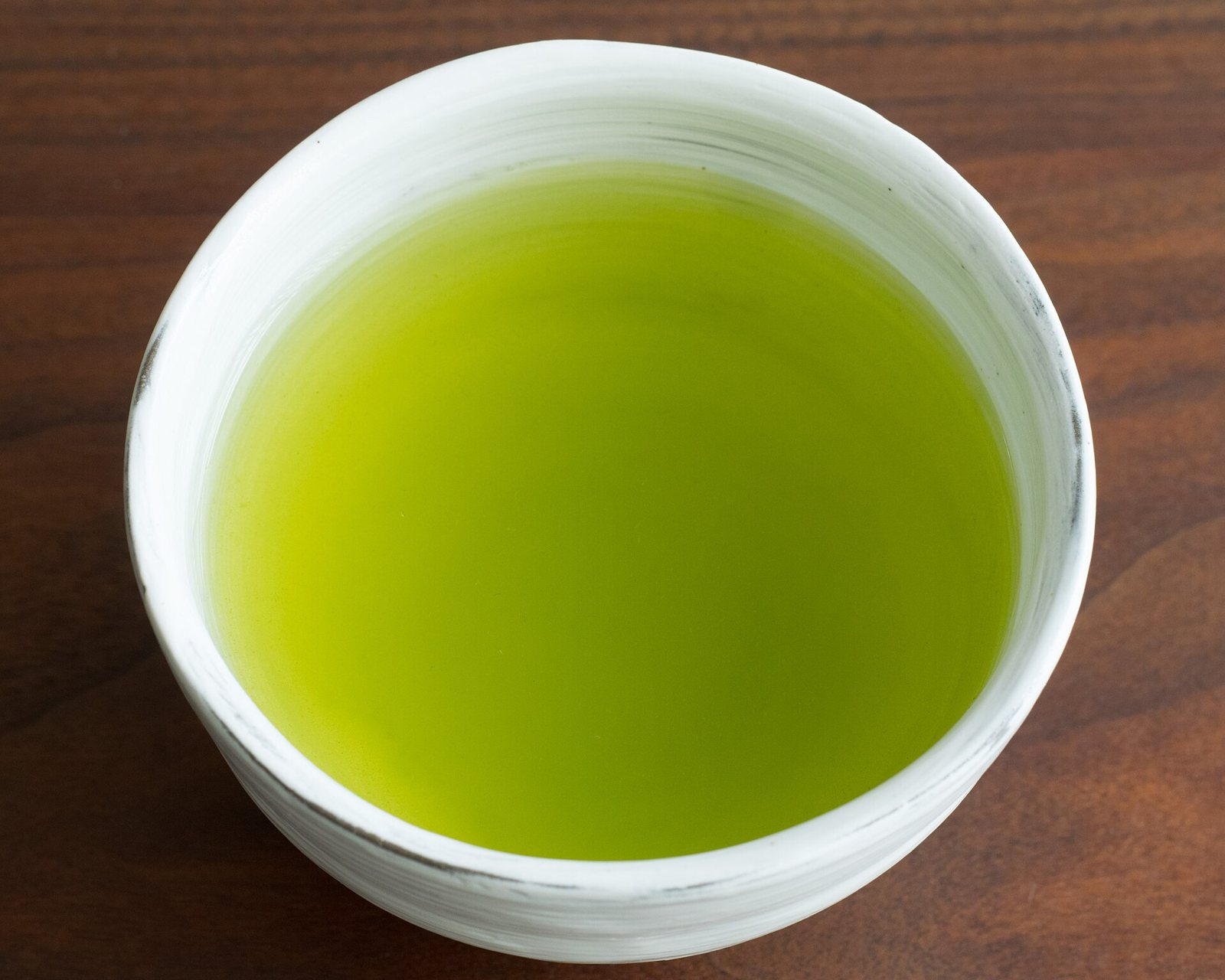 A white ceramic cup filled to near the brim with bright yellow-green sencha, seen from above on a dark wooden table.
